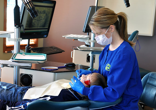 Dental professional in blue scrubs examining patient's teeth in orthodontic office treatment room.
