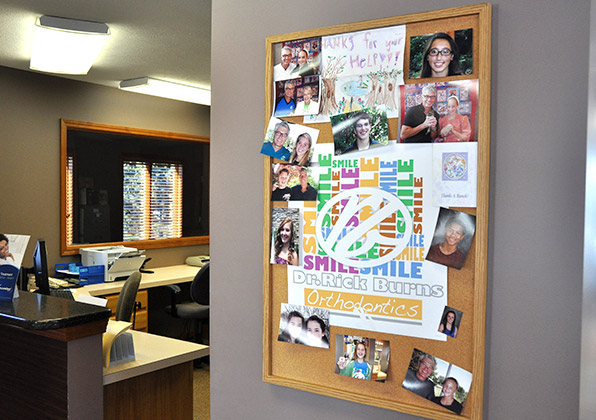 Orthodontic office bulletin board displaying patient photos, artwork, and certificates in wooden frame.