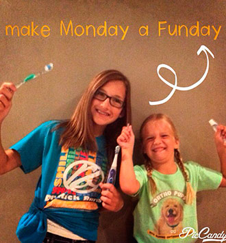 Two children smiling at camera against brown wall with "make Monday a Funday" text and drawn decorations.